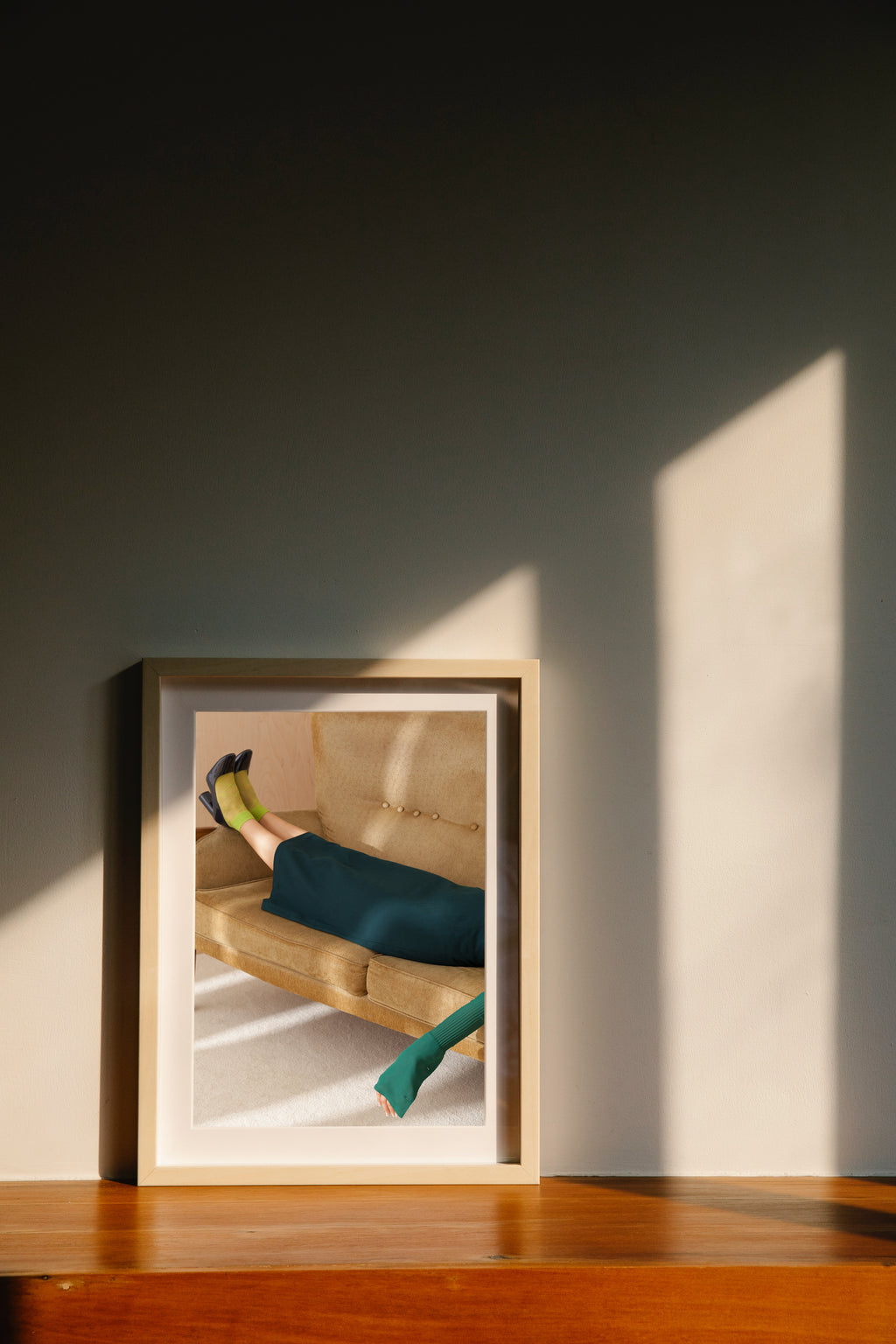 Wooden framed poster of a woman relaxing on a sofa, standing on the table in a sunlit room.